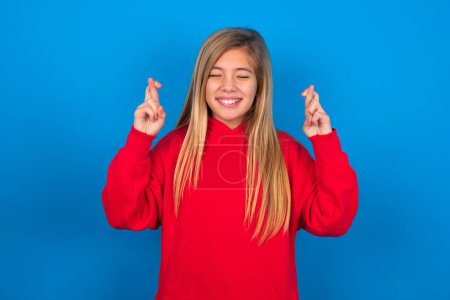 Photo for Joyful caucasian teen girl wearing red sweatshirt over blue background clenches teeth, raises fingers crossed, makes desirable wish, waits for good news, I have to win. - Royalty Free Image