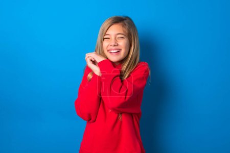 Photo for Dreamy caucasian teen girl wearing red sweatshirt over blue background with pleasant expression, closes eyes, keeps hands near face, thinks about something pleasant - Royalty Free Image