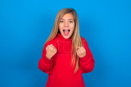 Téléchargez les photos : Belle adolescente caucasienne portant un sweat-shirt rouge sur fond bleu se réjouissant du succès et de la victoire serrant les poings avec joie étant heureuse d'atteindre son objectif et ses objectifs. Émotions positives, sentiments. - en image libre de droit