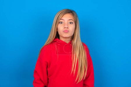 Foto de Encantadora chica adolescente caucásica con sudadera roja sobre fondo azul, mantiene los labios como va a besar a alguien - Imagen libre de derechos