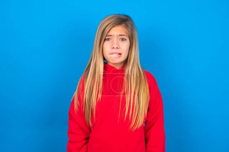 Photo for Beautiful caucasian teen girl wearing red sweatshirt over blue background being nervous and scared biting lips looking camera with impatient expression, pensive. - Royalty Free Image