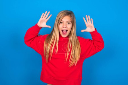 Photo for Beautiful caucasian teen girl wearing red sweatshirt over blue background looks with excitement at camera, keeps hands raised over head, notices something unexpected, reacts on sudden news. - Royalty Free Image