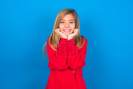 Téléchargez les photos : Dreamy caucasien adolescent fille portant sweat-shirt rouge sur fond bleu garde les mains pressées ensemble sous le menton, regarde avec une expression heureuse, a sourire aux dents. - en image libre de droit