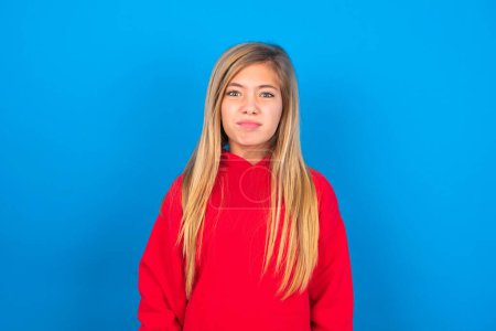 Téléchargez les photos : Caucasien adolescent fille portant sweat-shirt rouge fait grimace avec expression maladroite s'amuse seul, joue fou, tout en posant sur fond bleu studio - en image libre de droit