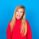 blonde caucasian teen girl wearing red sweatshirt with funny face showing tongue over blue studio background