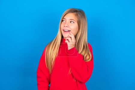 Photo for Beautiful caucasian teen girl wearing red sweatshirt over blue background with thoughtful expression, looks to the camera, keeps hand near face, biting a finger thinks about something pleasant. - Royalty Free Image