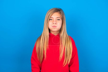 Photo for Smiling caucasian teen girl wearing red sweatshirt over blue background puffing cheeks with funny face. Mouth inflated with air, crazy expression. - Royalty Free Image