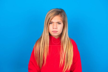 Téléchargez les photos : Belle caucasien adolescent fille portant rouge sweat-shirt sur fond bleu fronçant les sourcils être mécontent de quelque chose. - en image libre de droit