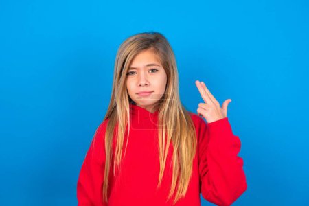Photo for Unhappy caucasian teen girl wearing red sweatshirt imitates gun shoot makes suicide gesture keeps two fingers on temples, posing over blue studio background - Royalty Free Image