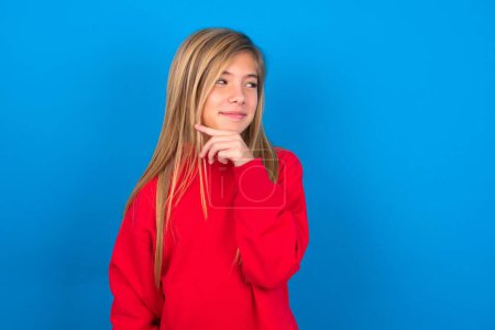 Photo for Dreamy caucasian teen girl wearing red sweatshirt over blue background with pleasant expression, looks sideways, keeps hand under chin, thinks about something pleasant. - Royalty Free Image