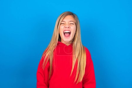 Stressful beautiful caucasian teen girl wearing red sweatshirt over blue background screams in panic, closes eyes in terror, finds out terrified news, can't believe it. 