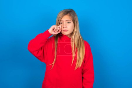 Foto de Infeliz caucásico adolescente chica usando rojo sudadera llorando mientras posando en cámara batiendo lágrimas con la mano sobre fondo azul - Imagen libre de derechos