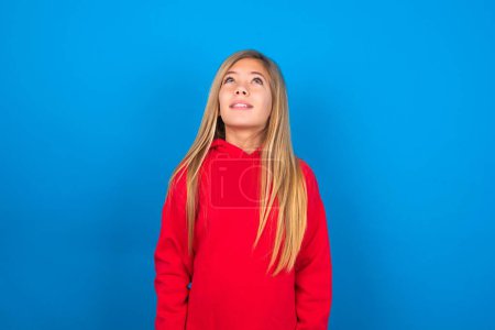 Téléchargez les photos : Blonde caucasien adolescent fille portant sweat-shirt rouge sur fond bleu regardant vers le haut comme elle voit quelque chose d'étrange. - en image libre de droit