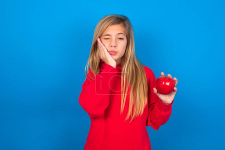 Foto de Hermosa chica adolescente caucásica con sudadera roja sobre fondo azul comiendo manzana, una mano en su mejilla - Imagen libre de derechos