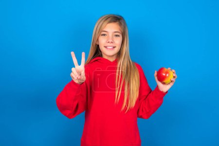 Photo for Beautiful caucasian teen girl with apple wearing red sweatshirt over blue background pointing up with fingers and showing number two while smiling confident and happy. - Royalty Free Image