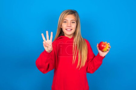 Photo for Beautiful caucasian teen girl with apple in hand wearing red sweatshirt showing with fingers number three while posing over blue background - Royalty Free Image