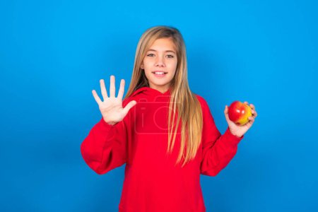 Téléchargez les photos : Belle adolescente caucasienne avec pomme à la main portant un sweat-shirt rouge montrant avec les doigts numéro cinq tout en posant sur fond bleu - en image libre de droit