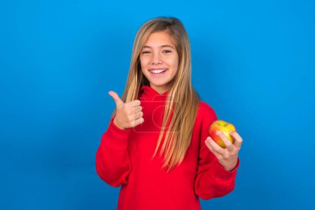 Foto de Hermosa chica adolescente caucásica con sudadera roja sobre fondo azul dando el pulgar hacia arriba gesto, recomendando comer manzana, estilo de vida saludable. - Imagen libre de derechos