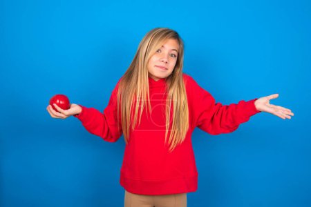 Photo for Puzzled and clueless caucasian teen girl wearing red sweatshirt with apple in hand and arms out, shrugging shoulders, saying: who cares, so what, I don't know. Negative human emotions. - Royalty Free Image