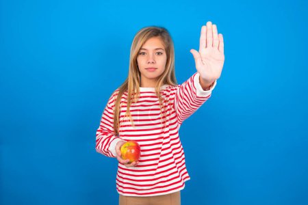 Foto de Adolescente niña con camisa a rayas sobre fondo azul haciendo gesto de parada con la palma de la mano. Expresión de advertencia con gesto negativo y serio en la cara. - Imagen libre de derechos