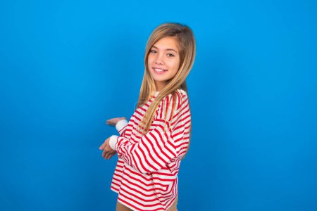 Foto de Adolescente chica vistiendo camisa a rayas sobre fondo azul invitando a entrar sonriendo natural con las manos abiertas. Señal de bienvenida. - Imagen libre de derechos