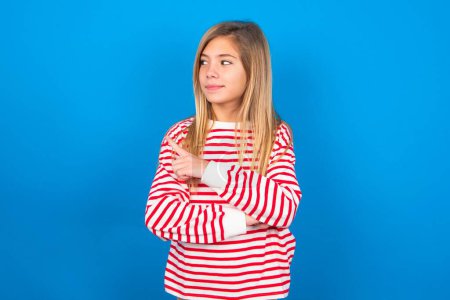Photo for Portrait of teen girl wearing striped shirt over blue background posing on camera with tricky look, presenting product with index finger. Advertisement concept. - Royalty Free Image