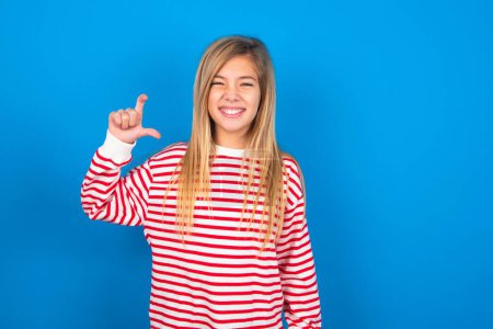 Foto de Adolescente chica vistiendo camisa a rayas sobre fondo azul sonriendo y haciendo un gesto con la mano de tamaño pequeño, símbolo de medida. - Imagen libre de derechos