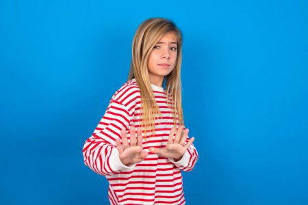 Photo for Afraid teen girl wearing striped shirt over blue background makes terrified expression and stop gesture with both hands saying: Stay there. Panic concept. - Royalty Free Image