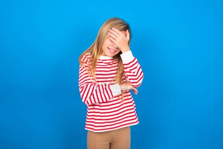 Téléchargez les photos : Adolescent fille portant rayé chemise sur fond bleu faire face geste tout en souriant étonné avec stupide situation sur fond bleu studio - en image libre de droit