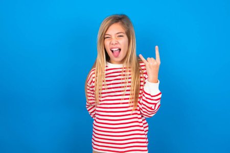 Téléchargez les photos : Portrait d'une adolescente folle portant une chemise rayée sur fond bleu montrant la langue cornes geste vers le haut, exprimant l'excitation d'être sur le concert du groupe. - en image libre de droit