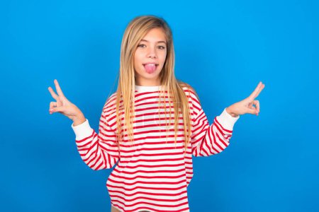Photo for Teen girl wearing striped shirt over blue background with optimistic smile, showing peace or victory gesture with both hands, looking friendly, posing over blue studio background - Royalty Free Image
