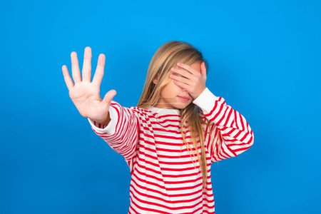 Photo for Teen girl wearing striped shirt over blue background covers eyes with palm and doing stop gesture, tries to hide. Don't look at me, I don't want to see, feels ashamed or scared. - Royalty Free Image