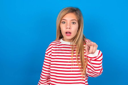 Photo for Shocked teen girl wearing striped shirt over blue background points front with index finger at camera and. Surprise and advertisement concept. - Royalty Free Image