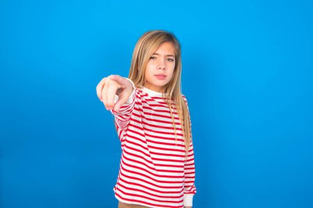 Téléchargez les photos : Joyeux adolescent fille portant une chemise rayée sur fond bleu indique joyeusement à vous, choisit de concourir, a une expression positive, fait un choix. - en image libre de droit