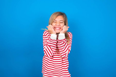 Photo for Portrait of teen girl wearing striped shirt over blue background being overwhelmed, expressing excitement and happiness with closed eyes and hands near face. - Royalty Free Image