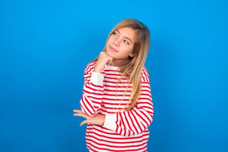 Photo for Face expressions and emotions. Thoughtful teen girl wearing striped shirt over blue background holding hand under his head, having doubtful look. - Royalty Free Image