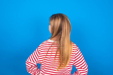 Photo for The back view of blonde teen girl wearing striped shirt over blue background over blue studio background - Royalty Free Image