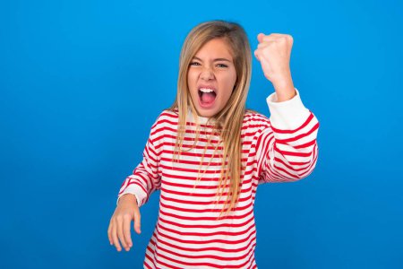 Foto de Chica adolescente feroz con camisa a rayas sobre fondo azul sosteniendo el puño en frente como si estuviera listo para luchar o desafiar, gritando y teniendo expresión agresiva en la cara. - Imagen libre de derechos