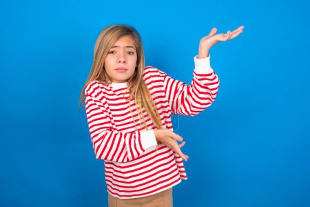 Téléchargez les photos : Belle adolescente caucasienne portant une chemise rayée sur fond bleu pointant de côté avec les deux mains montrant quelque chose d'étrange et disant : Je ne sais pas ce que c'est. Concept de publicité. - en image libre de droit