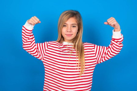 Photo for Waist up shot of teen girl wearing striped shirt over blue background raises arms to show muscles feels confident in victory, looks strong and independent, smiles positively at camera over blue studio background. Sport concept. - Royalty Free Image