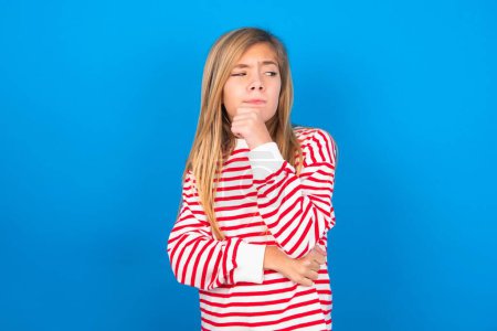 Téléchargez les photos : Adolescent fille portant chemise rayée sur fond bleu avec la main sous le menton et regardant de côté avec une expression douteuse et sceptique, suspect et doute. - en image libre de droit
