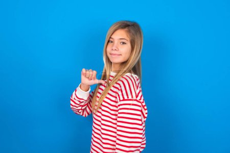 Closeup of cheerful teen girl wearing striped shirt over blue background looks joyful, satisfied and confident, points at himself with thumb.