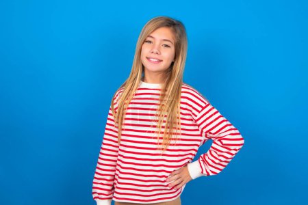 Photo for Studio shot of cheerful teen girl wearing striped shirt over blue background keeps hand on hip, smiles broadly. - Royalty Free Image