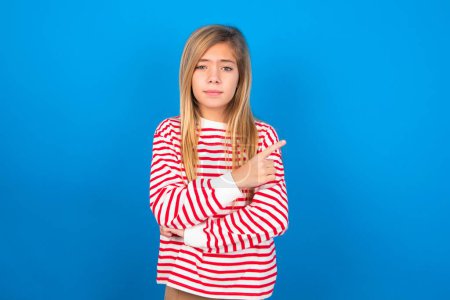 Photo for Teen girl wearing striped shirt over blue background smiling broadly at camera, pointing fingers away, showing something interesting and exciting. - Royalty Free Image