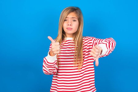 Foto de Adolescente chica vistiendo camisa rayada sobre fondo azul sentirse inseguro haciendo buena mala señal. Descontento y poco impresionado. - Imagen libre de derechos