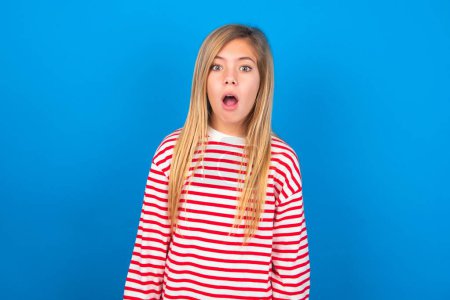 Photo for Emotional and attractive teen girl wearing striped shirt over blue background with opened mouth expresses great surprise and fright, stares at camera over blue studio background. Unexpected shocking news and human reaction. - Royalty Free Image