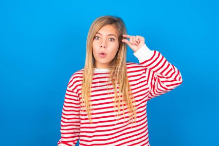 Photo for Shocked teen girl wearing striped shirt over blue background shows something little with hands, demonstrates size, opens mouth from surprise. Measurement concept. - Royalty Free Image