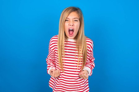 Photo for Joyful excited lucky teen girl wearing striped shirt over blue background cheering, celebrating success, screaming yes with clenched fists - Royalty Free Image
