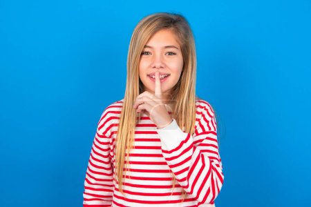 Foto de Sonriente chica adolescente con camisa a rayas sobre fondo azul hace gesto de silencio, sostiene el dedo delantero sobre los labios se esconde secreto. Silencio, por favor.. - Imagen libre de derechos