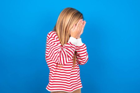 Téléchargez les photos : Triste adolescent fille portant une chemise rayée sur fond bleu couvrant le visage avec les mains et pleurant. - en image libre de droit